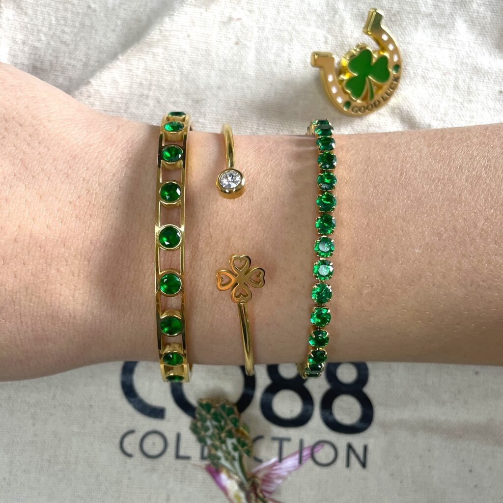 Composition de bracelets verts Co88 collection. Les trois bracelets sont en acier inoxydable doré. Il y a un bracelet rivière, un jonc avec un trèfle et un diamant et le troisième un jonc qui se ferme avec des zircons verts foncés
